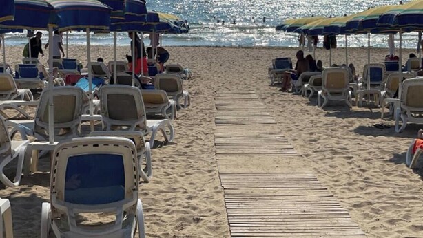 Une plage bordée de rangées de chaises longues et de parasols, une promenade en bois menant à l’eau, et des personnes à l’arrière-plan.