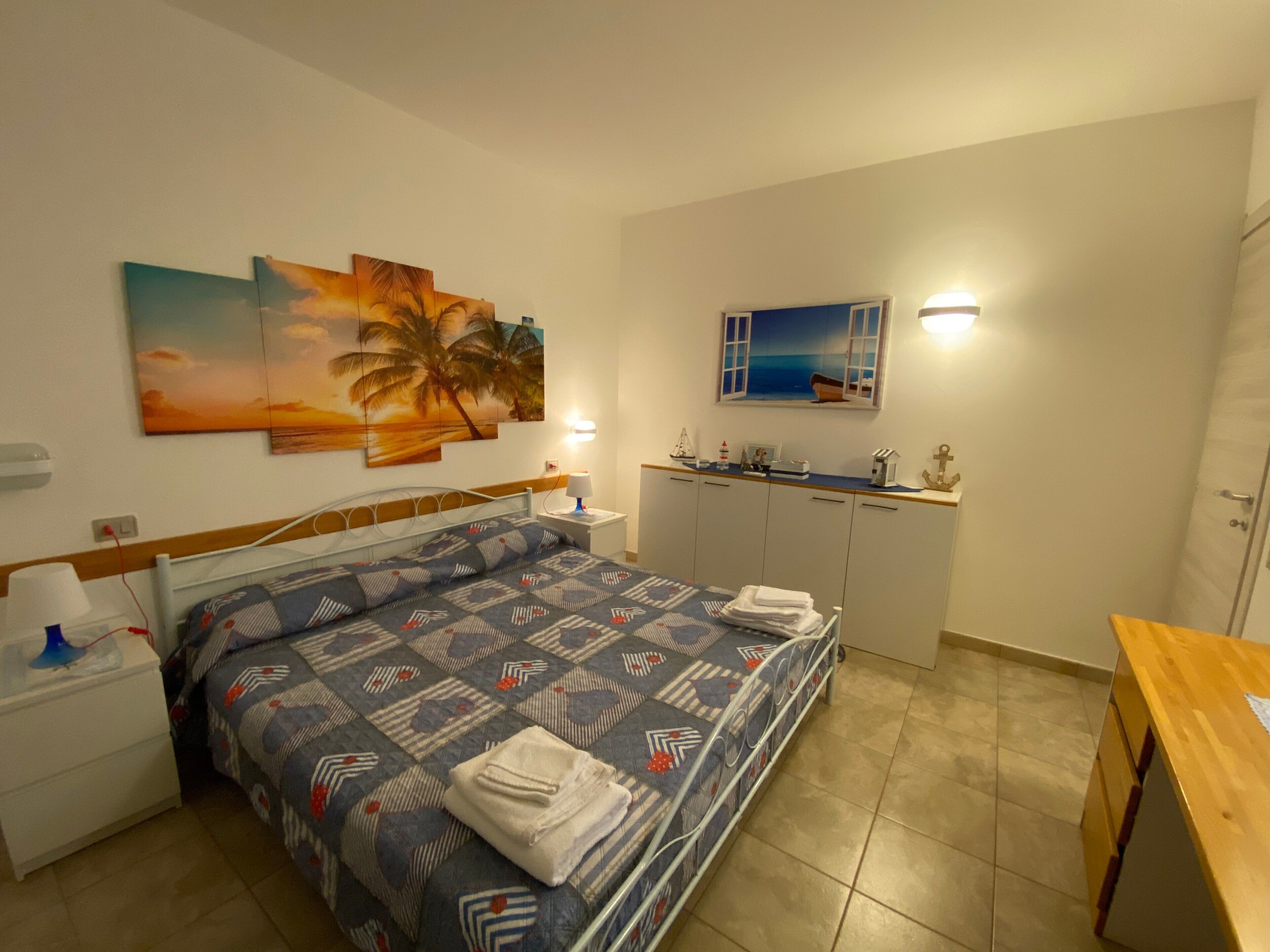 Une chambre à coucher comprenant un lit, une table de chevet, un bureau, une armoire et un tableau représentant un coucher de soleil avec des palmiers.