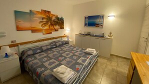 Une chambre à coucher comprenant un lit, une table de chevet, un bureau, une armoire et un tableau représentant un coucher de soleil avec des palmiers.