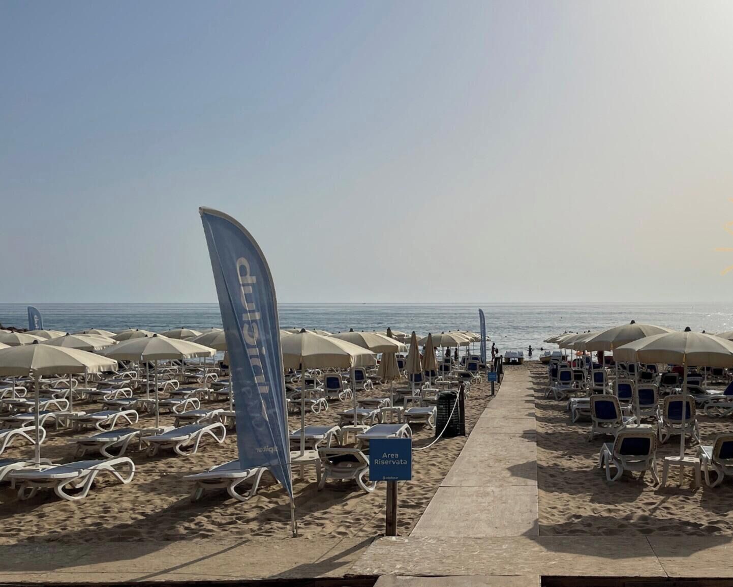Une plage bordée de rangées de chaises longues et de parasols, avec un drapeau et un panneau indiquant une zone réservée à un groupe spécifique.