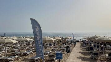 Une plage bordée de rangées de chaises longues et de parasols, avec un drapeau et un panneau indiquant une zone réservée à un groupe spécifique.
