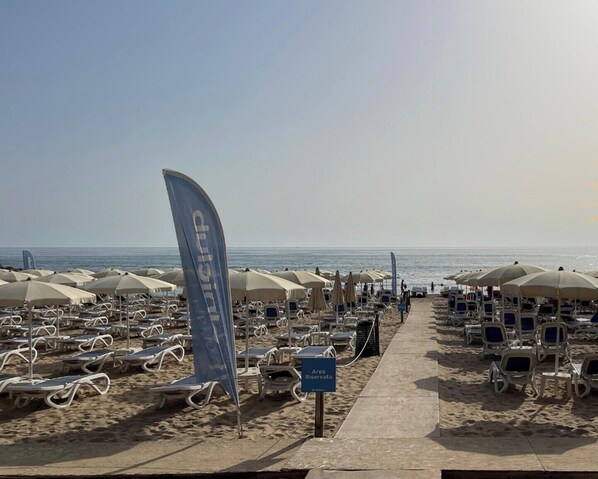 A beach with rows of sun loungers and umbrellas, a flag, and a sign indicating an area reserved for a specific group. - Holiday Home "Kastalia - Blu" with Sea View, Shared Pool and Wi-Fi (Ragusa)
