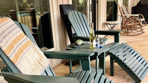A covered patio area with green Adirondack chairs, a small table with a bottle and glass, and a wooden deck.