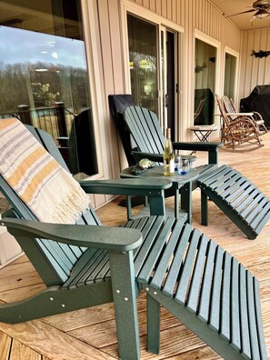 Un patio cubierto con sillas Adirondack verdes, una mesita con una botella y un vaso, y una terraza de madera.