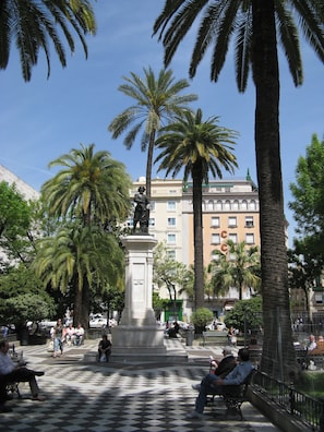 A public square with a statue, palm trees, and a building with balconies in the background.