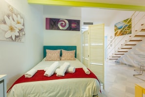 A hotel room with a bed, pillows, a red blanket, and towels. There is a painting on the wall and a staircase leading to another room.