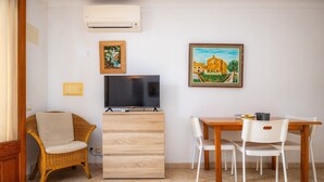 Une pièce comprenant un téléviseur posé sur un meuble en bois, une chaise en osier, une table à manger avec des chaises, et deux tableaux encadrés accrochés au mur.