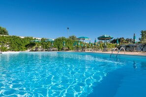 Une piscine d’un bleu éclatant, bordée de chaises longues et d’ombrelles, et entourée de verdure.