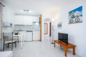 A modern kitchen with white cabinets, a dining area with a table and chairs, a TV on a wooden stand, and a painting of a bicycle on the wall.