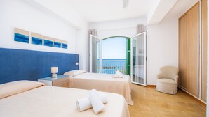 A hotel room with two beds, a chair, a nightstand, and a view of the sea through a glass door.