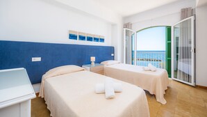 A hotel room with two beds, a small table, a nightstand, and a view of the sea through a glass door.