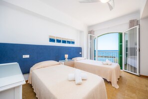 A hotel room with two beds, a small table, a nightstand, and a view of the sea through a glass door.