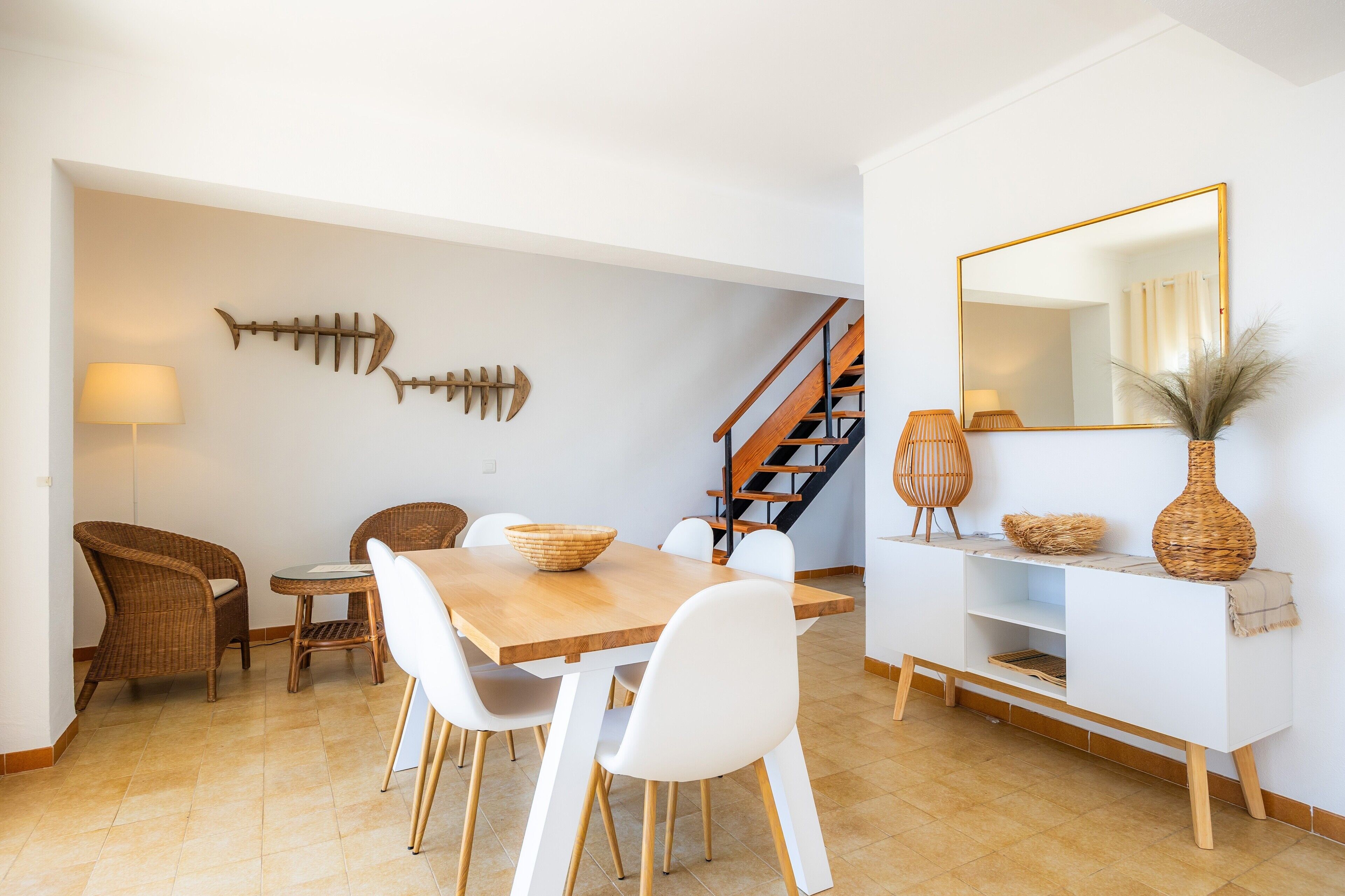 Zona pranzo con tavolo in legno, sedie bianche, poltrone in vimini, una decorazione a forma di pesce, una scala, un tavolino consolle bianco con oggetti decorativi e un grande specchio.
