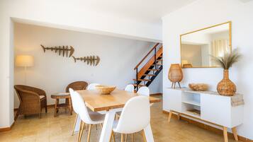 Zona pranzo con tavolo in legno, sedie bianche, poltrone in vimini, una decorazione a forma di pesce, una scala, un tavolino consolle bianco con oggetti decorativi e un grande specchio.