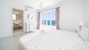 A bedroom with a bed, a dresser, a mirror, a window with curtains, and a view of the sea.