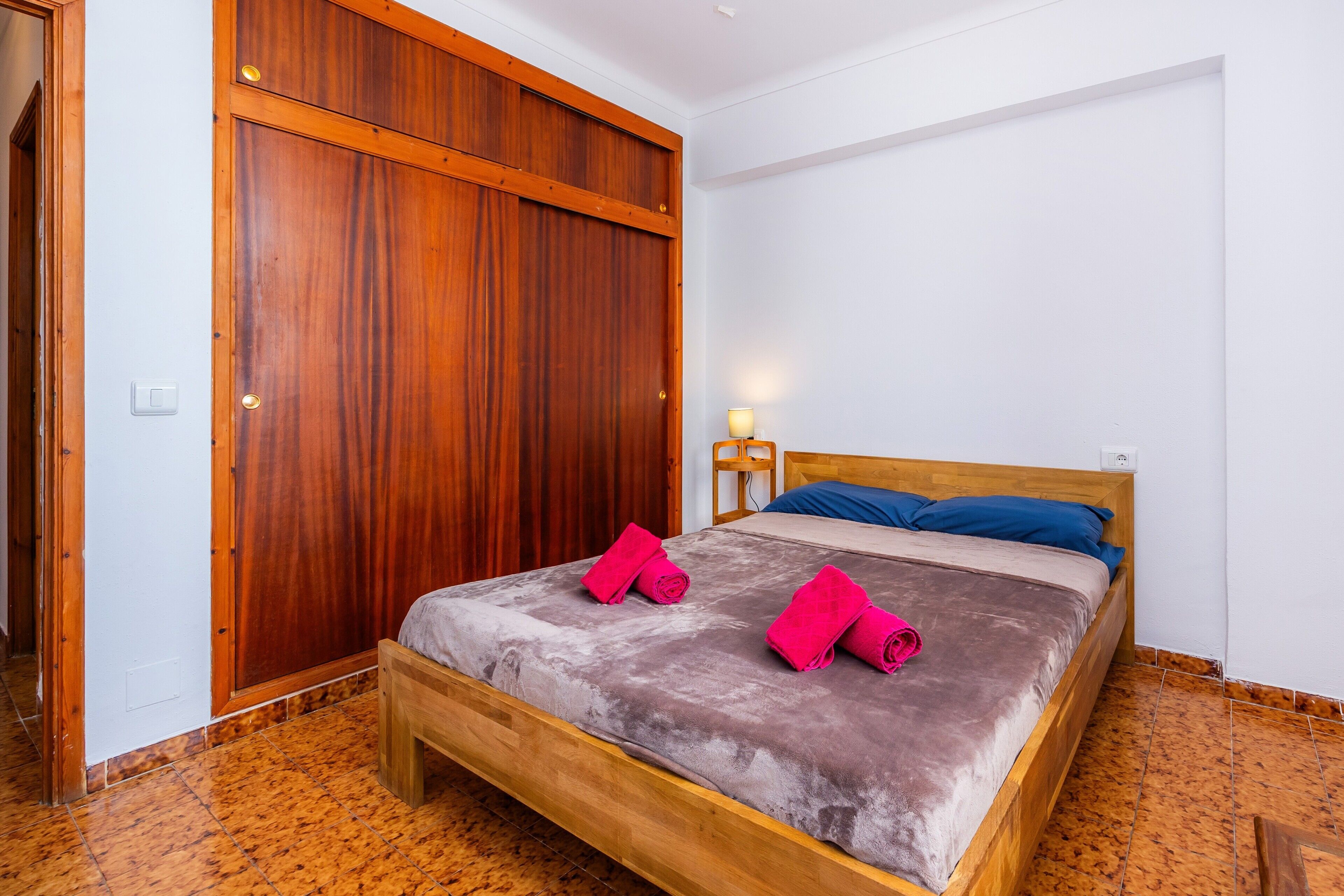 Una habitación con una cama de madera, un armario grande, una mesita de noche con lámpara y suelo de baldosas.