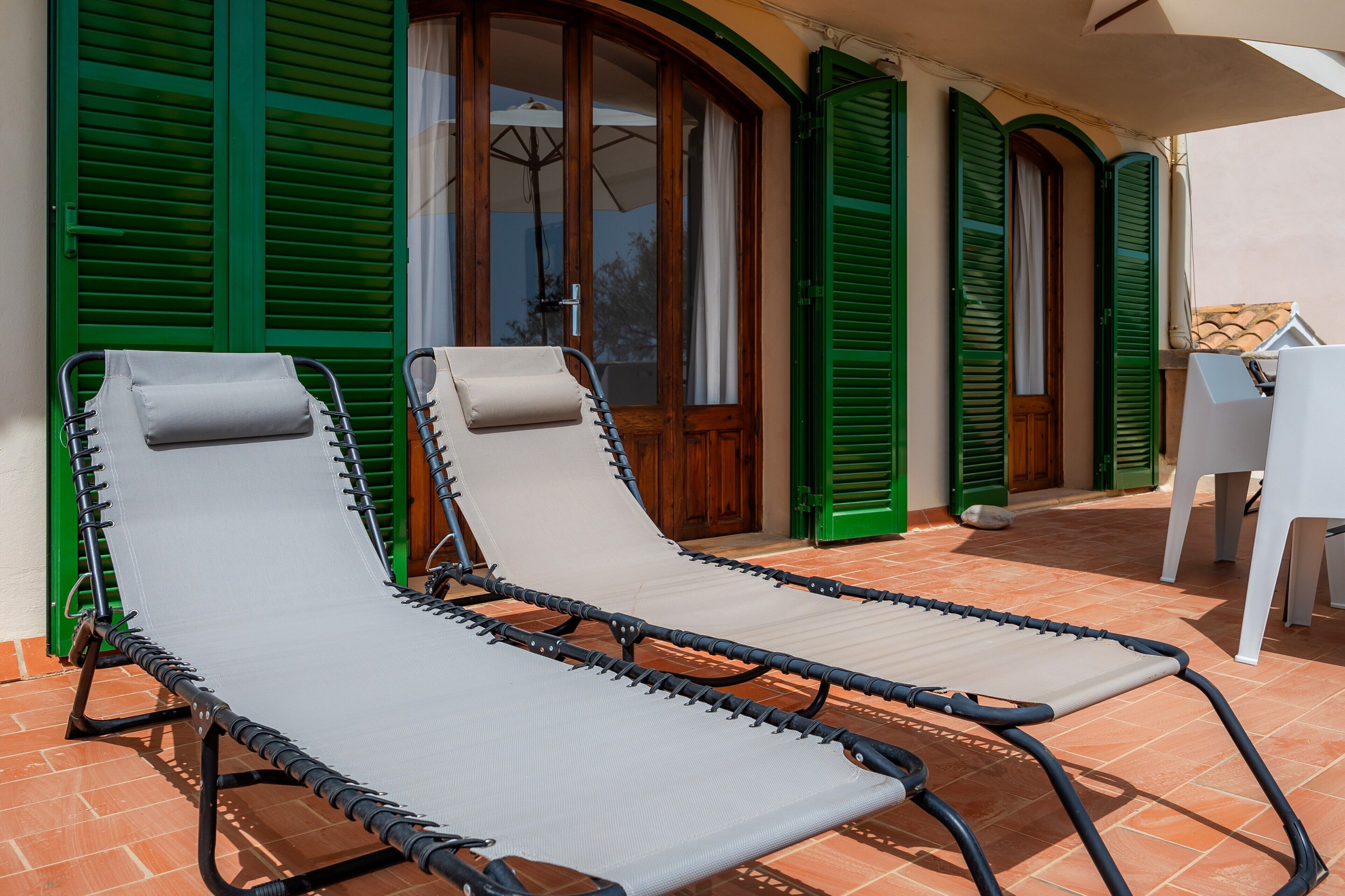 Dos sillones de relax en un patio con baldosas, contraventanas verdes y una mesa blanca.