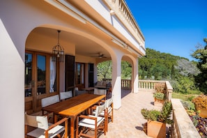 A covered outdoor dining area with a wooden table and chairs, a ceiling fan, and a view of a lush garden and hillside.