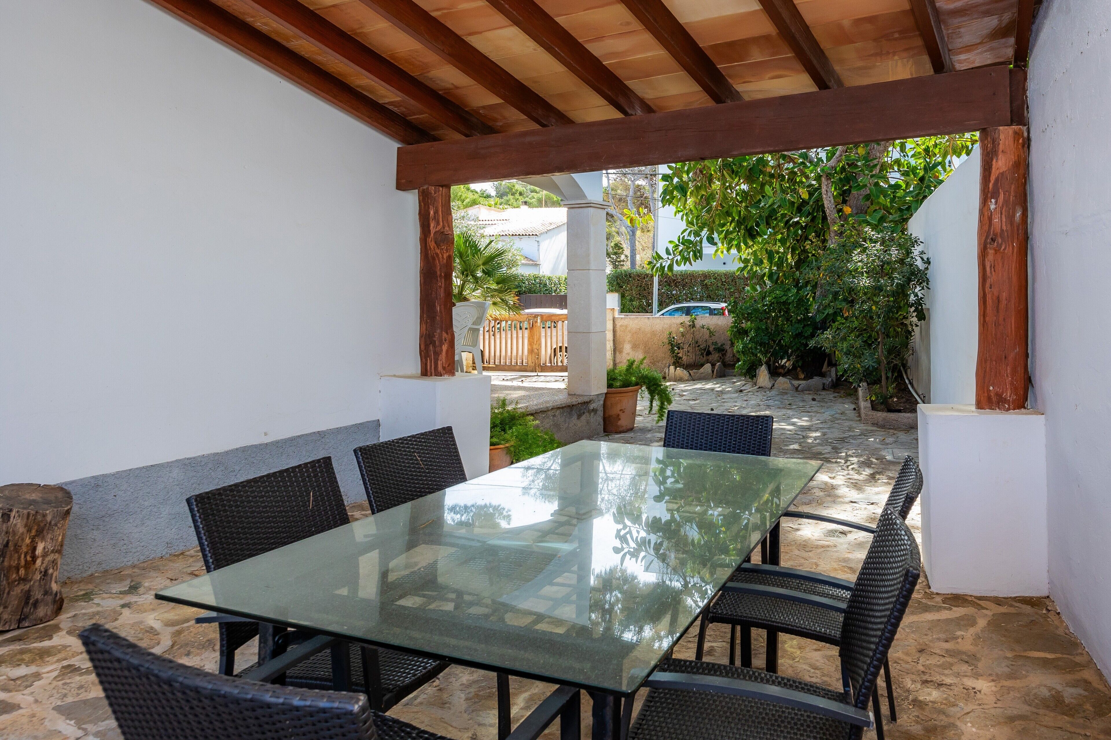 An outdoor patio with a glass table and wicker chairs, surrounded by white walls and a wooden pergola.