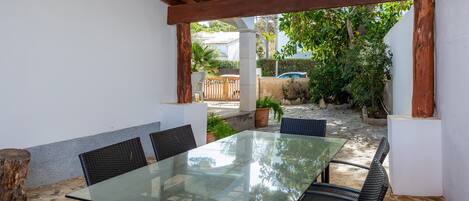 An outdoor patio with a glass table and wicker chairs, surrounded by white walls and a wooden pergola.