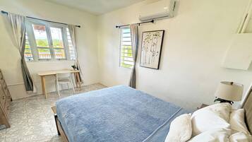 Un dormitorio con cama, escritorio y silla. Hay una ventana con cortinas, aire acondicionado y un cuadro en la pared.