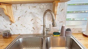 A kitchen with a stainless steel sink, a wooden shelf above, and a window with a view of greenery. - Beach House with Pool in Playa Santa - Casa Perla (Guánica)