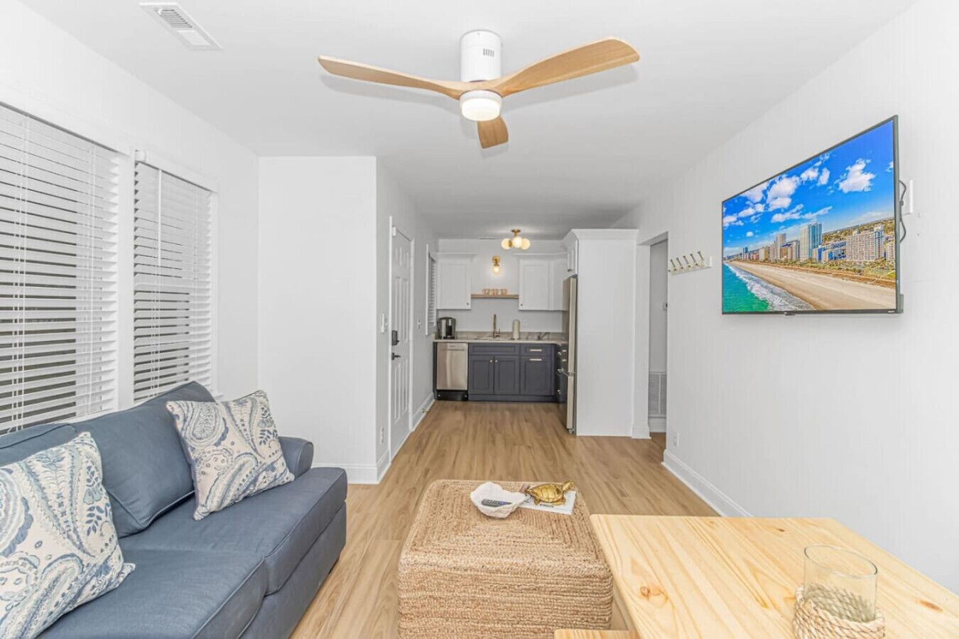A modern living room with a grey sofa, a wooden coffee table, and a ceiling fan. A kitchen is visible in the background with white cabinets and a view of the city.