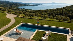 Una piscina con vistas a un lago y montañas, rodeada por un césped cuidado y algunas sillas.