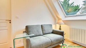 A small, modern living room with a grey sofa, a glass coffee table, and a window with a view of trees. - Ker Zen - Locmariaquer (LOCMARIAQUER)
