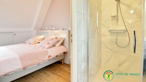 A bedroom with a bed, a nightstand, and a bedside table with a lamp. There is a shower with a glass door and a handheld showerhead. The room has a white ceiling and walls. - Ker Zen - Locmariaquer (LOCMARIAQUER)