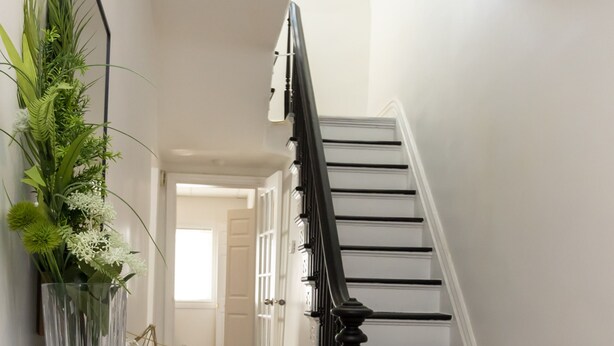 Una escalera con barandillas negras y peldaños blancos que conduce a una puerta. En la base, una mesa blanca con un florero y una cesta de mimbre.