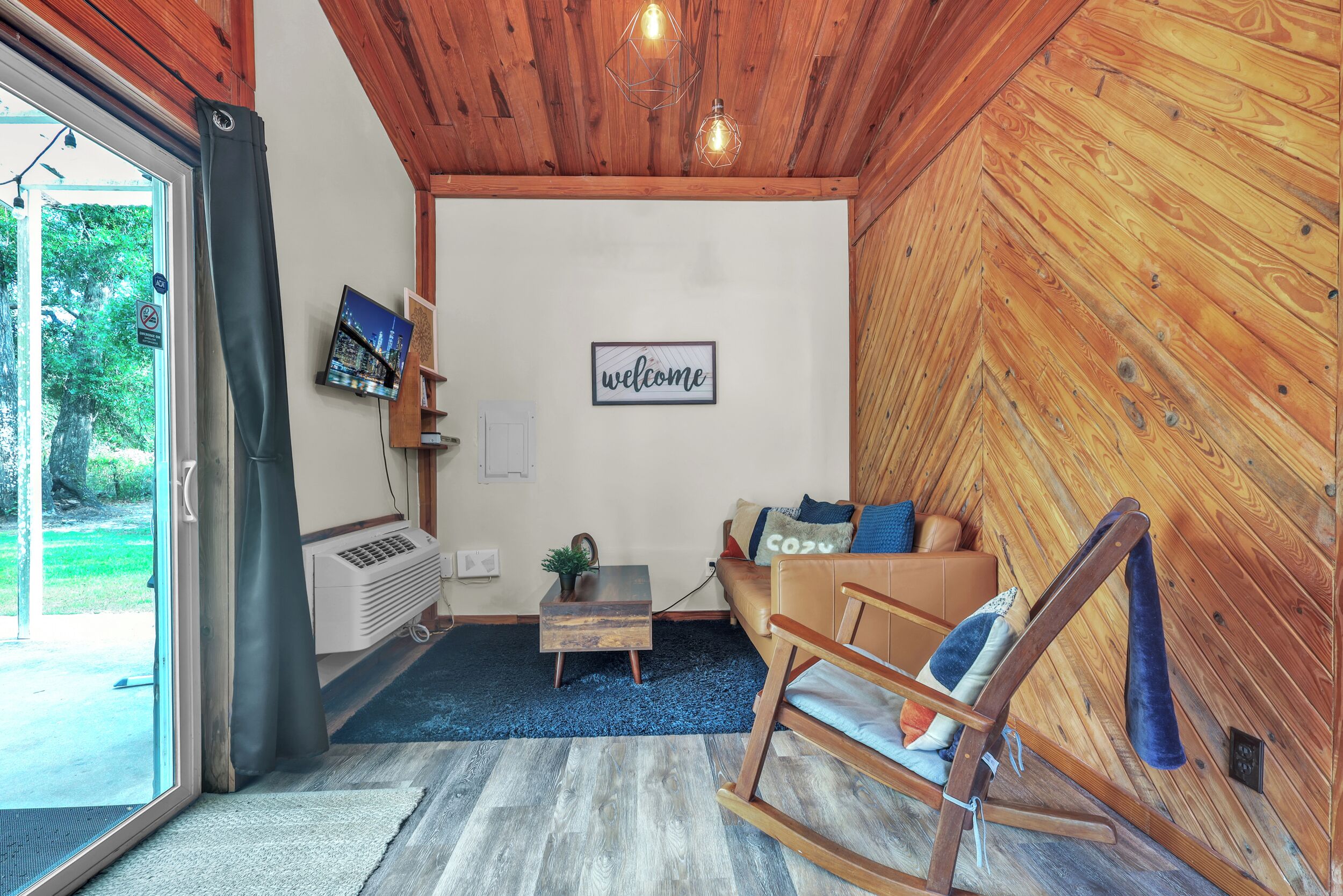 A cozy room with a wooden ceiling, a rocking chair, a sofa, a small table, and a TV on the wall.