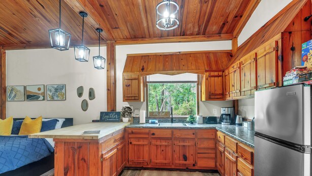 A kitchen with wooden cabinets, a stainless steel refrigerator, and a window overlooking greenery. - The Loft  Private Cabin Near Hulaween & Spirit of the Suwannee (Live Oak)
