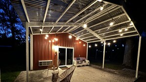 A covered outdoor patio with a wooden wall, a sliding glass door, and string lights. - The Loft  Private Cabin Near Hulaween & Spirit of the Suwannee (Live Oak)