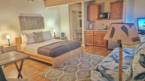 Ein Schlafzimmer mit Bett, Nachttischen, Schreibtisch, Stuhl, einer Couch, einem gemusterten Teppich, einer Küchenzeile mit Schränken und einer Mikrowelle sowie einem Fernseher.
