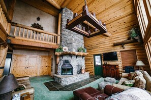A log cabin interior with a stone fireplace, wooden walls, a green carpet, and a wooden staircase.