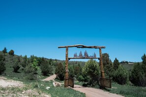 A rustic wooden archway with a metal gate, surrounded by a natural landscape with trees and a dirt path.