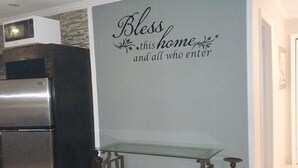 A kitchen with a stainless steel refrigerator, a microwave, and a glass-top table with a bench. A decorative wall sign reads "Bless this home and all who enter." - 
5 mins away from public beach and 3 min from grocery store! (The Forest)