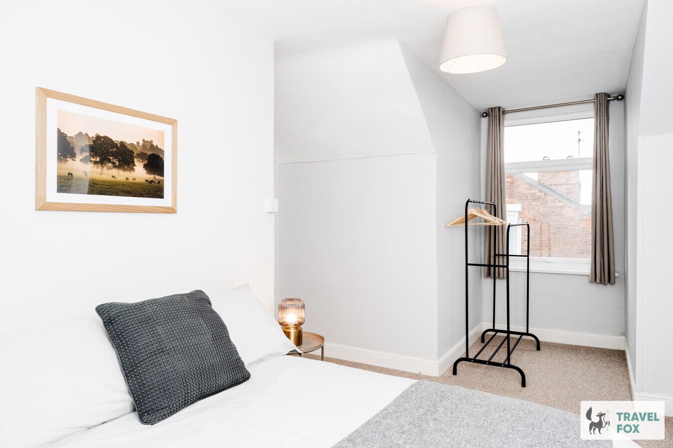 Une chambre à coucher avec un lit, une table de chevet avec une lampe, un porte-manteau mural, une fenêtre avec des rideaux et un tableau encadré au mur.