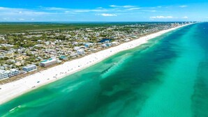 Vista aérea de un pueblo costero con una extensa playa de arena, aguas turquesas cristalinas y una zona residencial densa con edificios a lo largo de la costa.