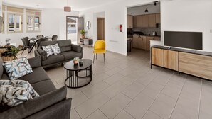 A modern living room with a sofa, coffee table, and a TV. There is a dining area with chairs and a table. The kitchen is visible with cabinets and a stove. - Villa Allegra: Private Pool & Modern Comfort (Protaras)