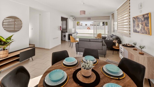 A modern dining area with a round table set for four, a sofa, and a kitchen in the background. - Villa Allegra: Private Pool & Modern Comfort (Protaras)
