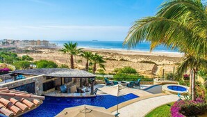 Un espace piscine offrant une vue sur la plage et l’océan, entouré de palmiers et de chaises longues.