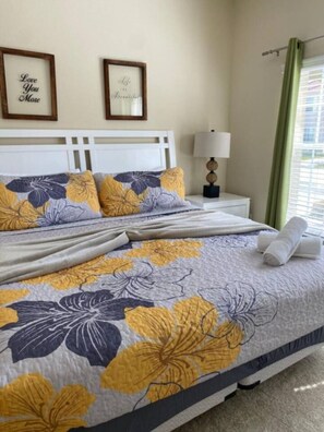 A neatly made bed with floral bedding, a white headboard, two framed inspirational quotes, a bedside lamp, and a window with green curtains. - Disney escape with private pool resort amenities (Kissimmee)