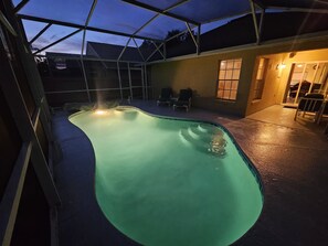 A swimming pool with a curved edge, surrounded by a covered patio area with chairs and a table. - Magical Getaway Near Disney & Universal – Family & Pet Friendly! (Kissimmee)