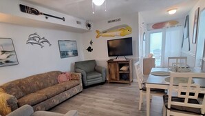 Un salon avec un canapé gris, un fauteuil gris, une table à manger en bois avec des chaises blanches, un ventilateur de plafond et un téléviseur.