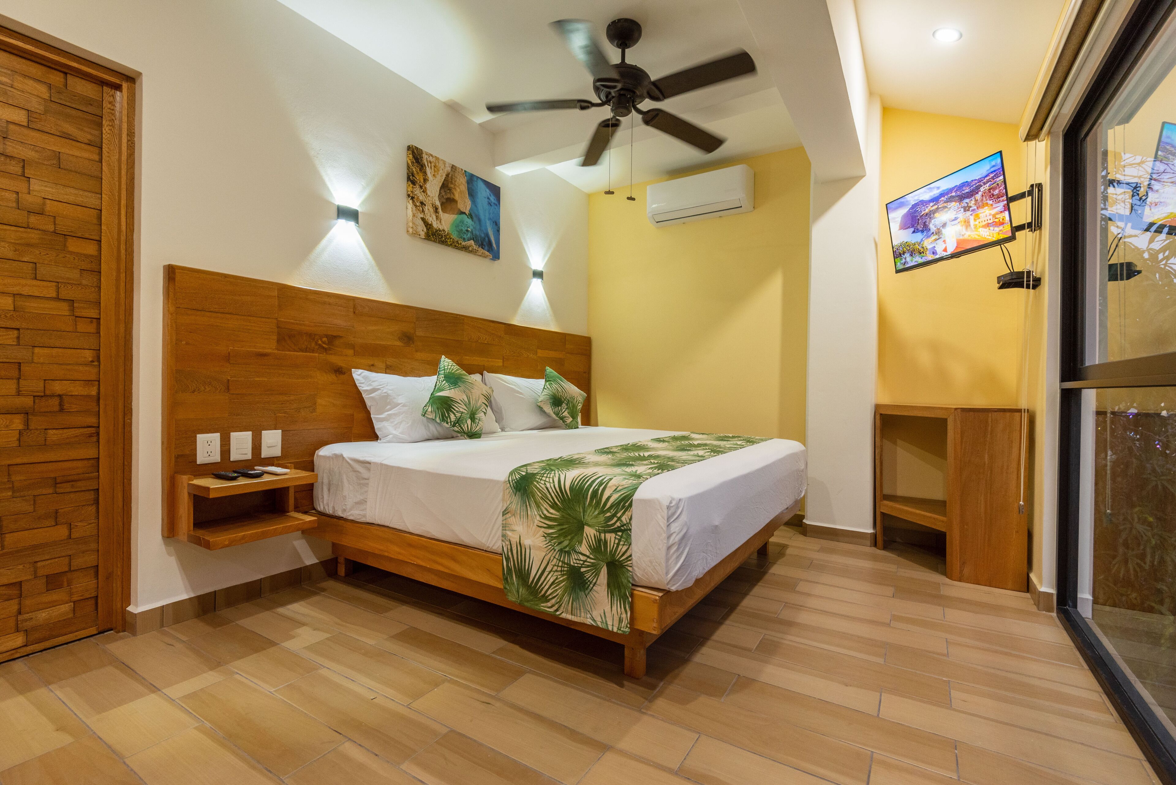 Quarto com cabeceira de madeira, cama com colcha branca e manta estampada com folhas de palmeira, mesa de cabeceira com controle remoto, ventilador de teto, ar condicionado e TV fixada na parede.
