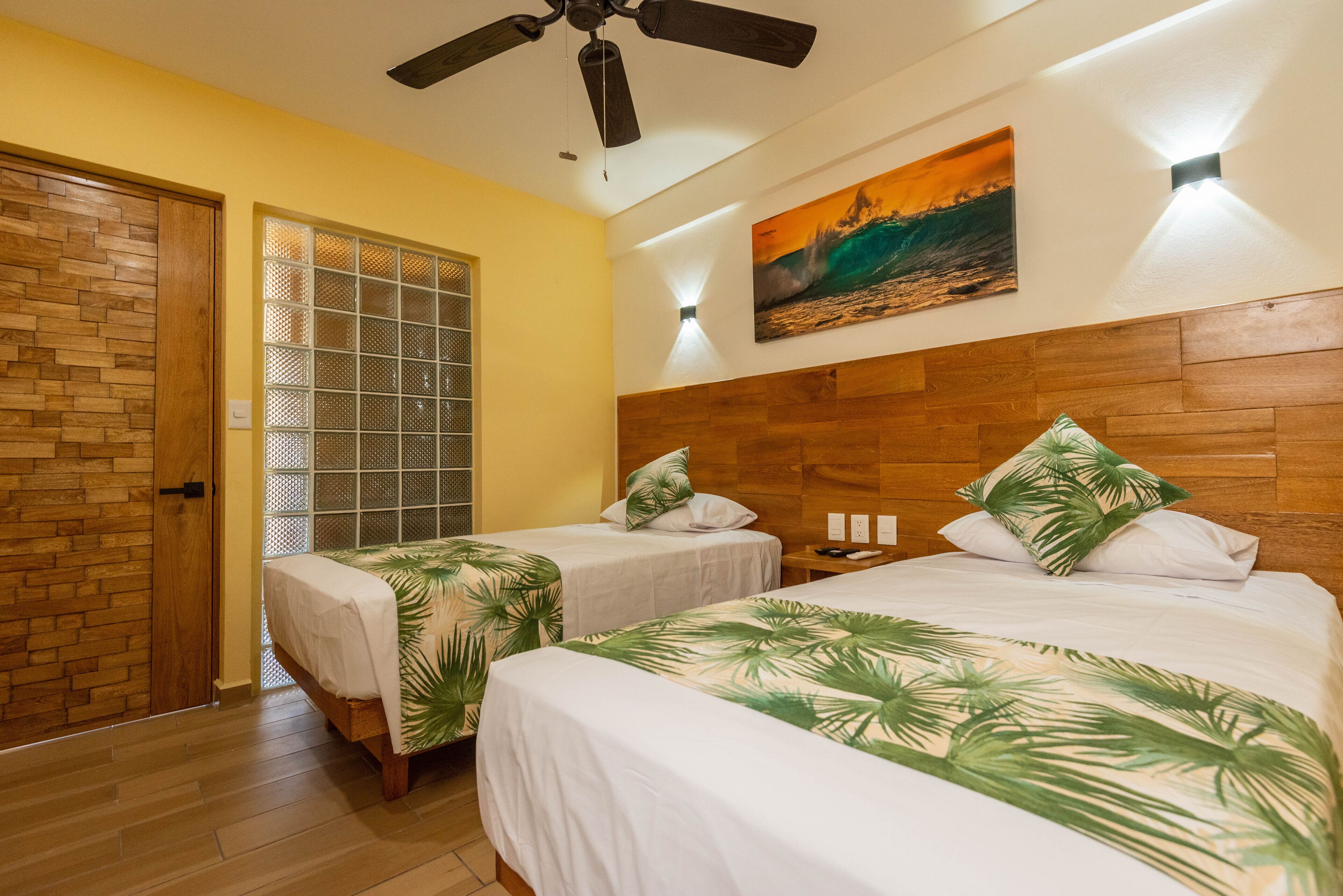 A hotel room with two beds, a wooden headboard, a ceiling fan, a beach painting, and a window with a decorative screen.