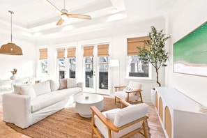 A modern living room with a white sofa, wooden chairs, a round coffee table, and a large window with blinds.