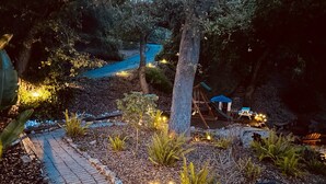 Un sendero en el jardín con un columpio, un área de juegos, rodeado de árboles e iluminado con luces colgantes.